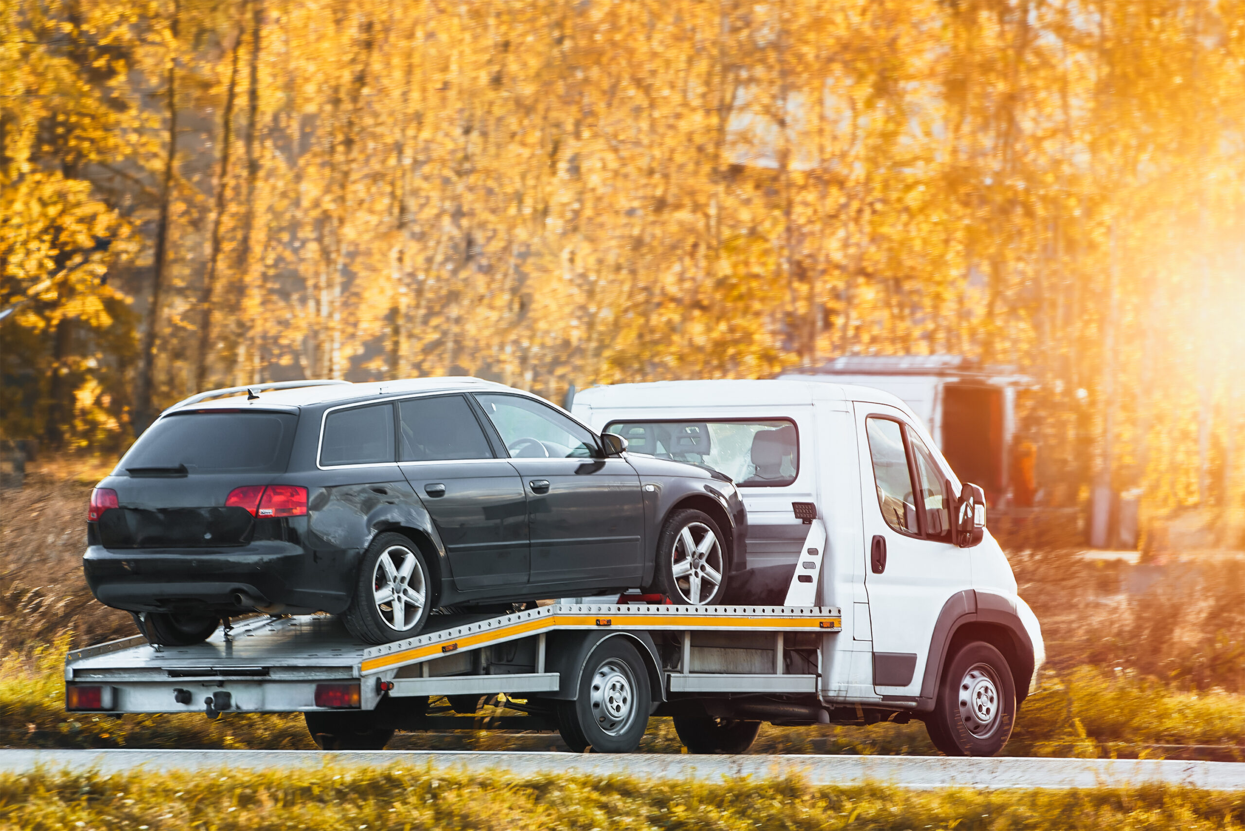 a roadside assistance service on a country road. A tow truck is carrying a car that has been involved in a traffic accident and has a broken engine